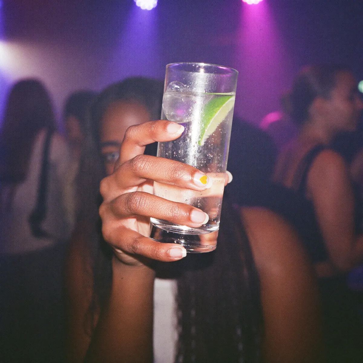 Una mano sujeta un vaso alto en un club con luces violeta; en la uña, la pegatina de Spy Nails.