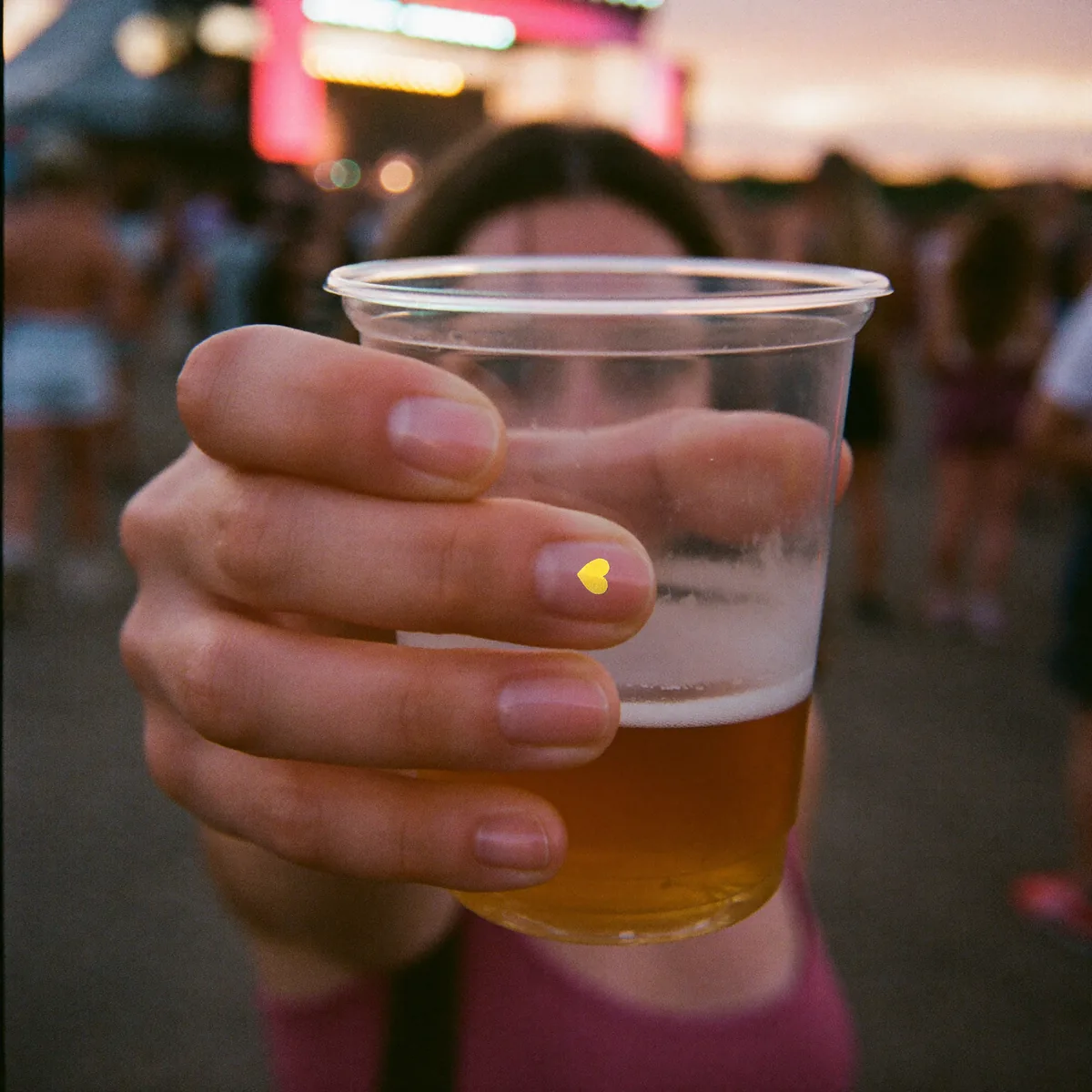 Una mano sujeta un vaso de cerveza en un festival al atardecer; en la uña, la pegatina de Spy Nails.