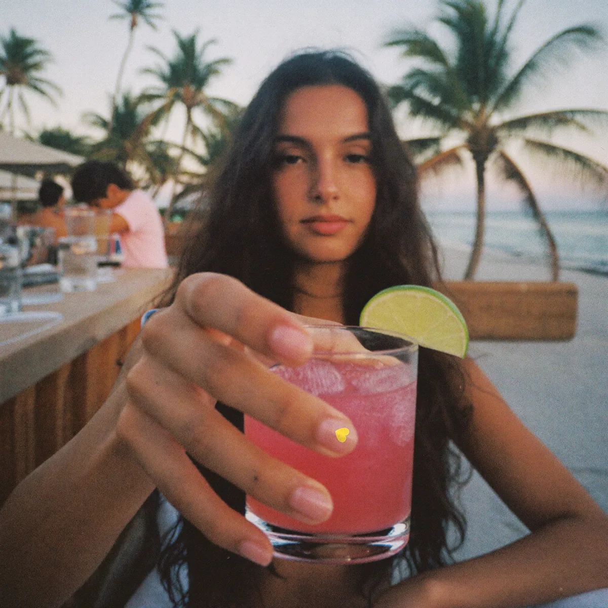 Una chica sujeta un cóctel rosa en un chiringuito de playa; en la uña, la pegatina de Spy Nails.